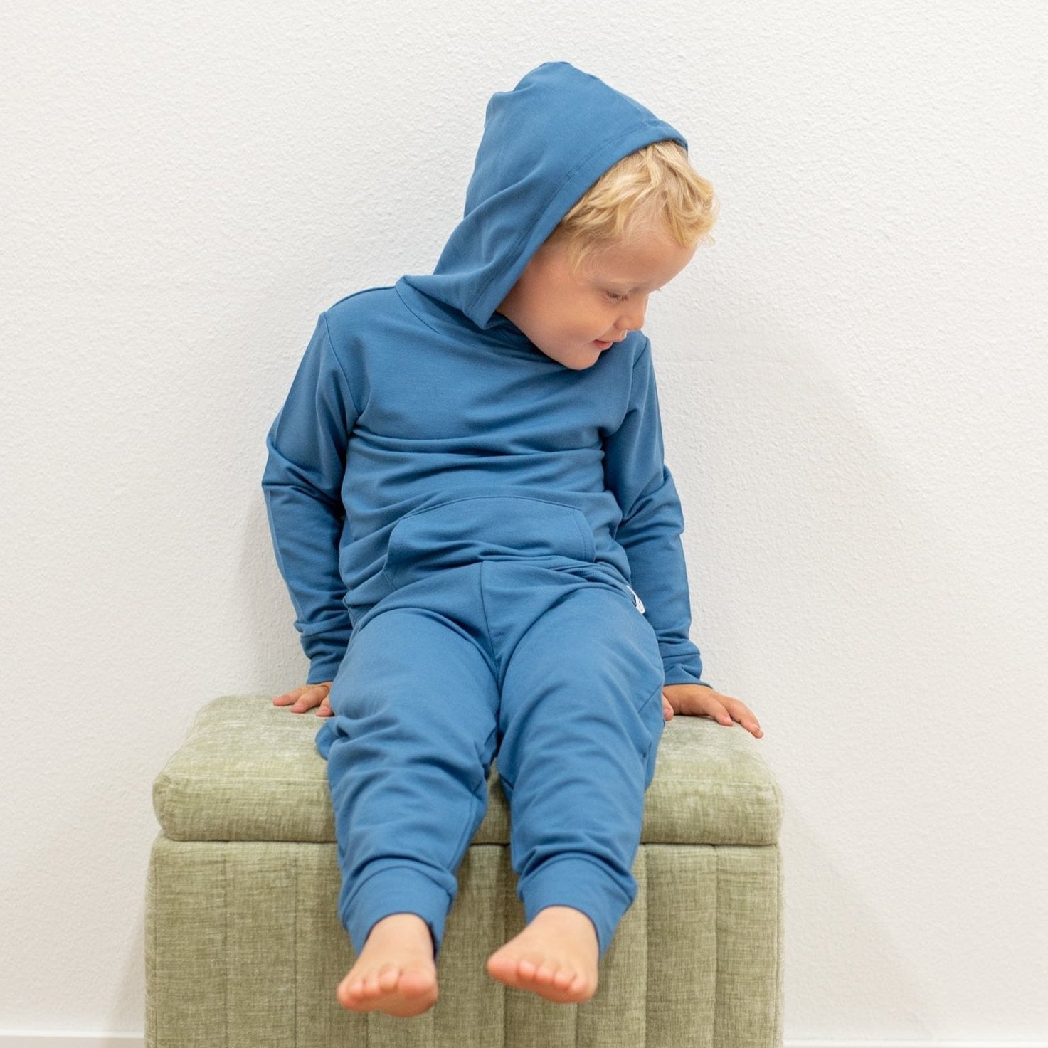 Child wearing a blue hoodie and pants sitting on a green ottoman against a white wall.