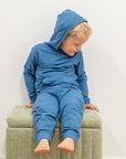 Child wearing a blue hoodie and pants sitting on a green ottoman against a white wall.