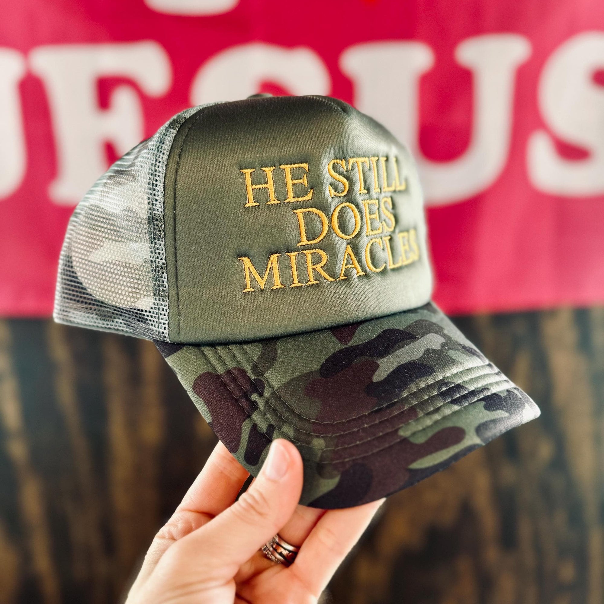 Camouflage cap with 'He Still Does Miracles' text held by a hand, with a blurred background.