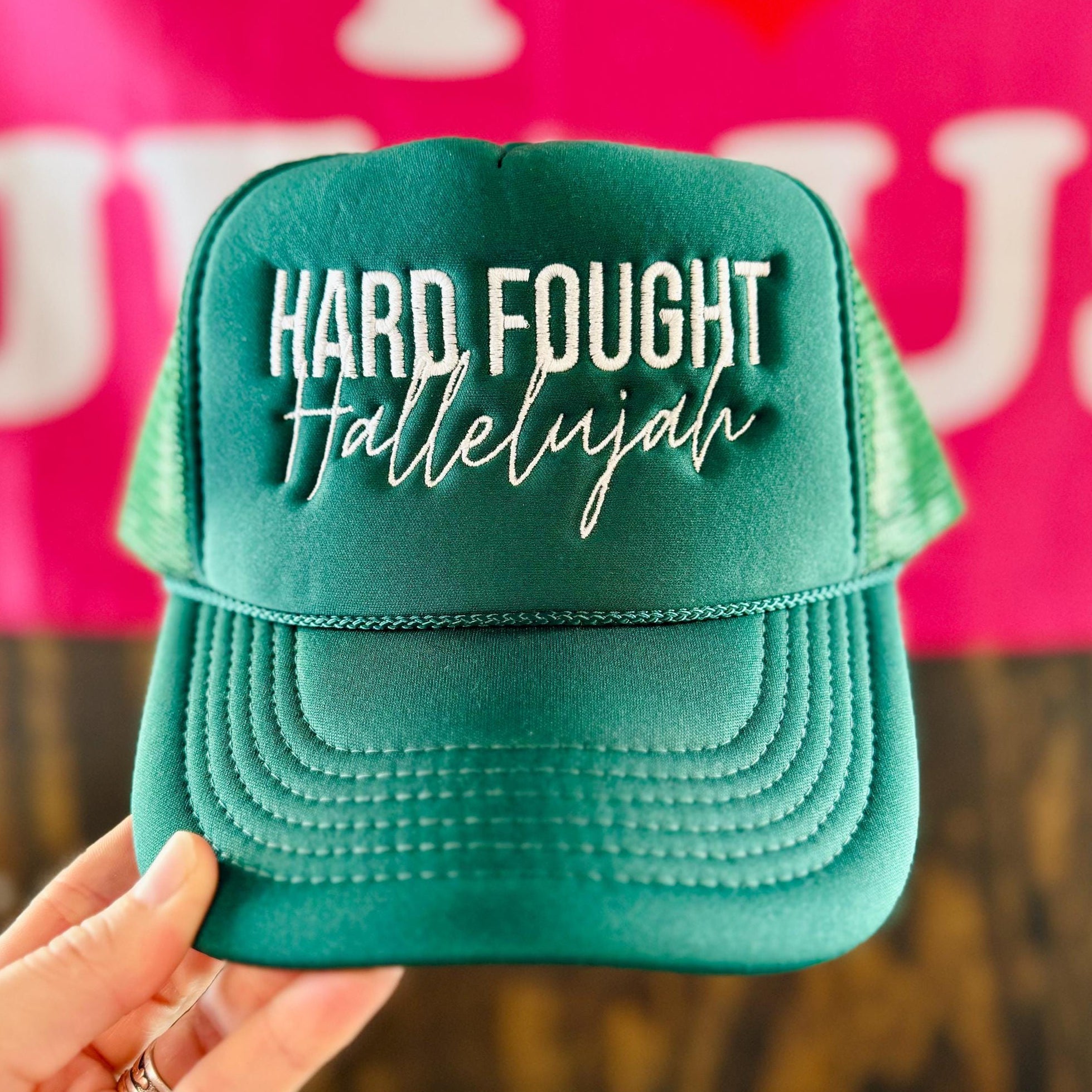 Teal cap with 'Hard Fought Hallelujah' text held in front of a pink background with 'I ❤️ Jesus' text.