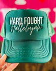 Teal cap with 'Hard Fought Hallelujah' text held in front of a pink background with 'I ❤️ Jesus' text.