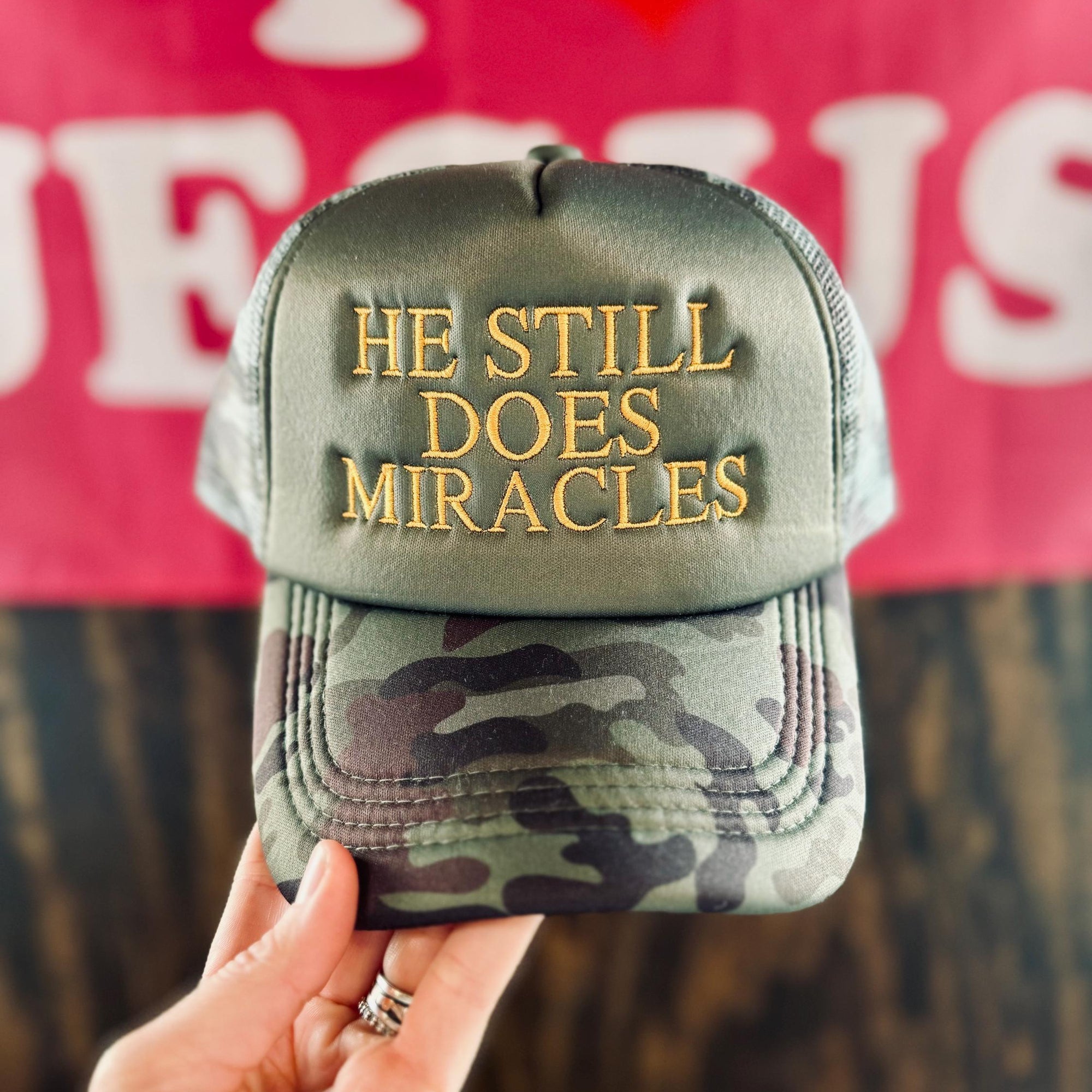 Camouflage cap with 'He Still Does Miracles' text held by a hand against a blurred background