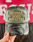 Camouflage cap with 'He Still Does Miracles' text held by a hand against a blurred background