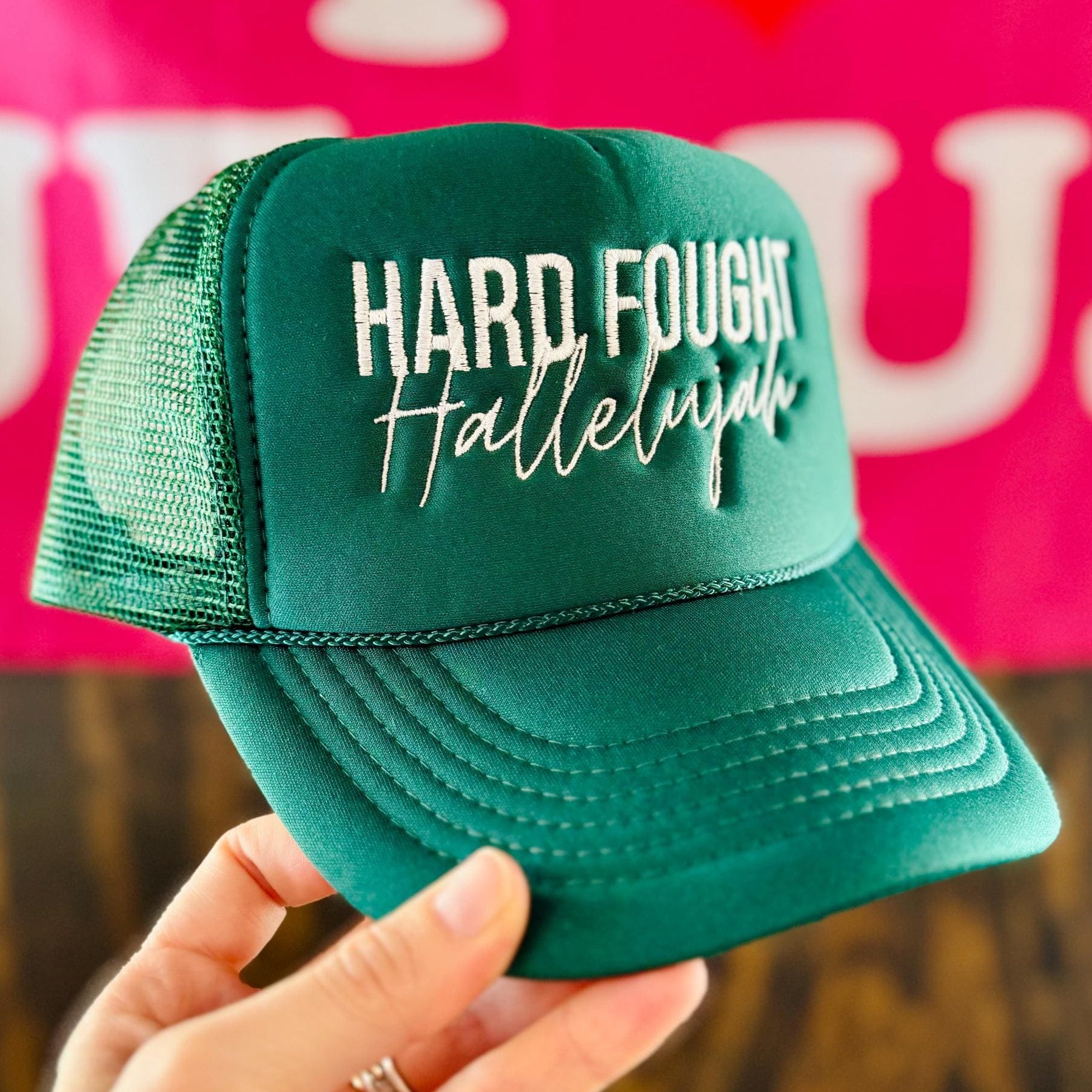 Green cap with 'Hard Fought Hallelujah' text held in front of a pink background with 'I ❤️ LUCY' text.