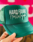Green cap with 'Hard Fought Hallelujah' text held in front of a pink background with 'I ❤️ LUCY' text.