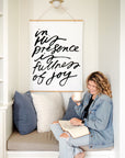 In His Presence scripture wall art with black script on a white background above a reading nook where a woman holds a mug and an open book.