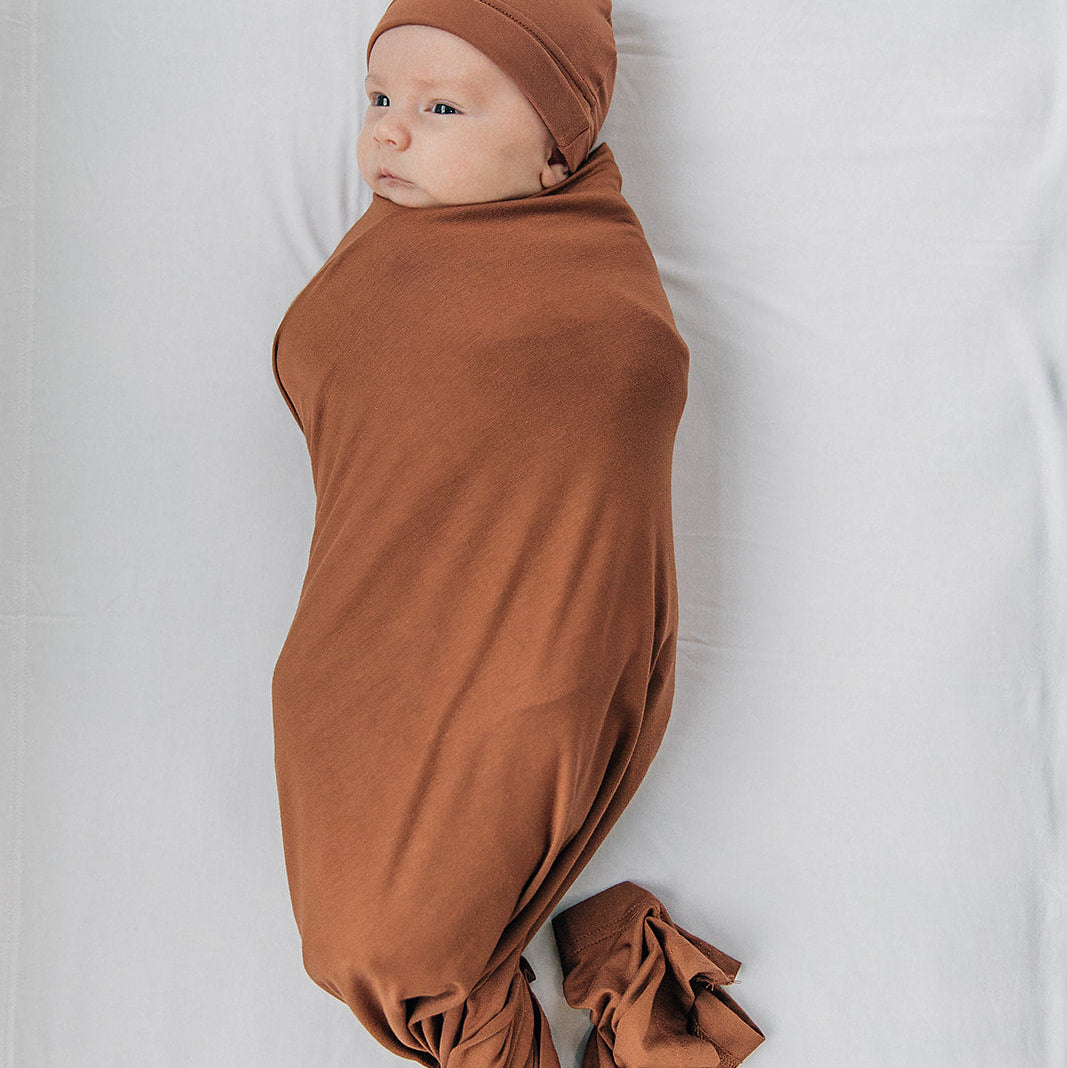Newborn baby swaddled in brown fabric with a matching headband on a white background