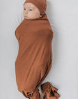 Newborn baby swaddled in brown fabric with a matching headband on a white background