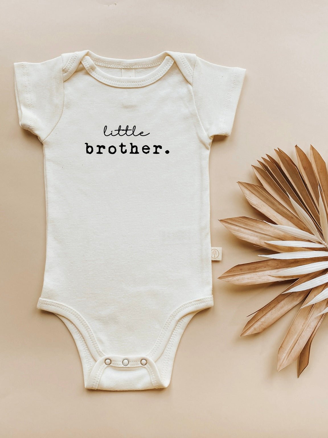 Little Brother organic cotton baby bodysuit laid flat beside dried foliage on a neutral background