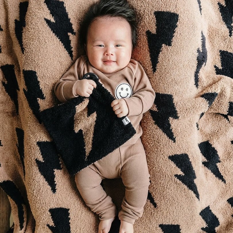 Baby in a brown outfit holding a black and white blanket on a patterned blanket
