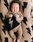 Baby in a brown outfit holding a black and white blanket on a patterned blanket