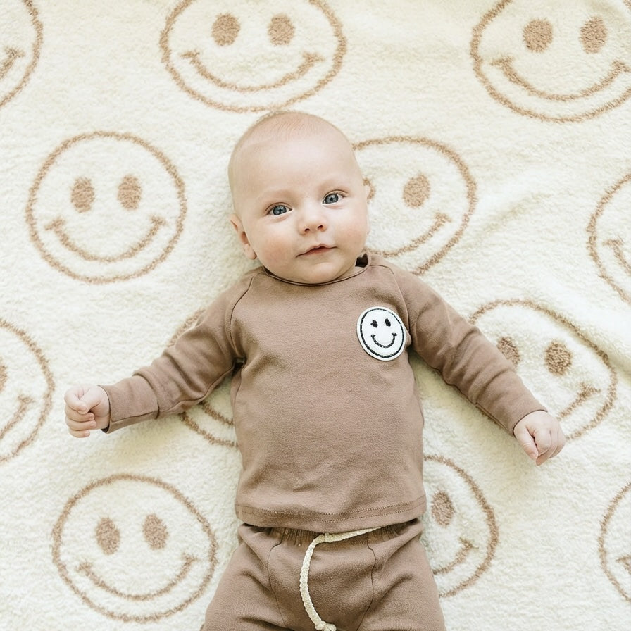Baby wearing a brown outfit with a smiley face on a beige rug with smiley face patterns