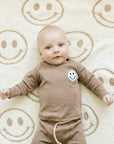 Baby wearing a brown outfit with a smiley face on a beige rug with smiley face patterns