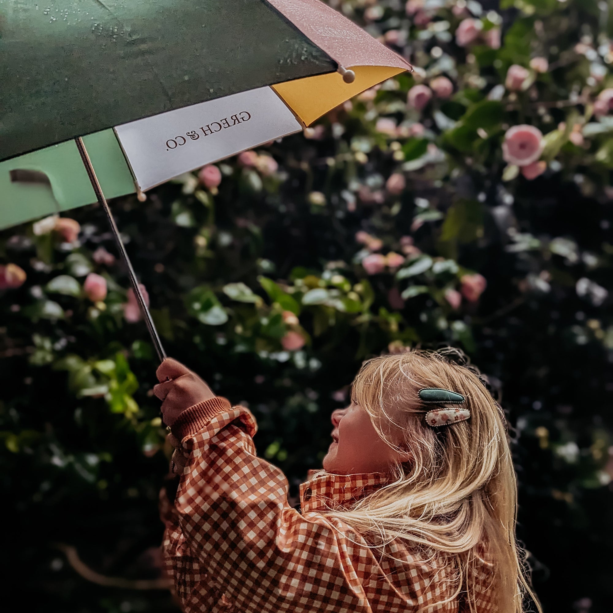 Person holding a green umbrella with a visible brand logo, standing in front of a floral background.
